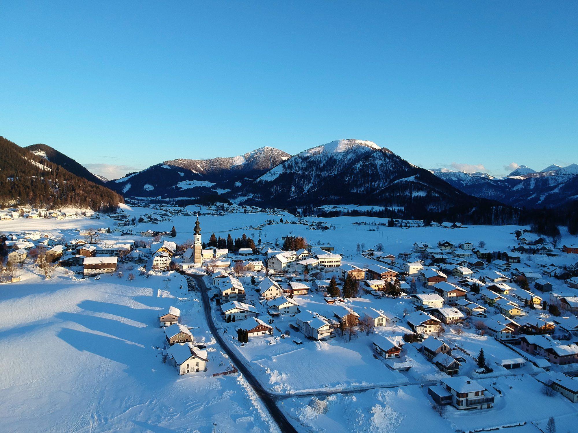 Faistenau im Winter von oben: verschneite Häuser rund um die Kirche, dahinter die Berge